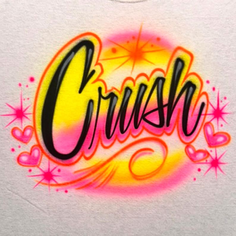 May include: White t-shirt with an airbrushed design. The word "Crush" is written in black, outlined in yellow and orange, with a pink and yellow gradient background. Pink hearts and starbursts surround the word.