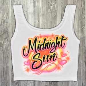 May include: White crop top featuring the words "Midnight Sun" in a vibrant airbrushed style. The design incorporates pink hearts, orange and yellow hues, and starbursts. The top has a scoop neck and is sleeveless.