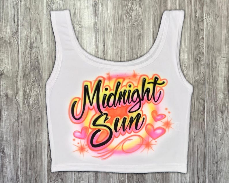 May include: White crop top featuring the words "Midnight Sun" in a vibrant airbrushed style. The design incorporates pink hearts, orange and yellow hues, and starbursts. The top has a scoop neck and is sleeveless.