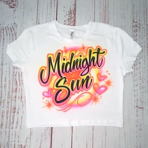 May include: White crop top with the words "Midnight Sun" in a colorful airbrushed design. The design includes pink hearts and orange starbursts, creating a vibrant and eye-catching graphic.