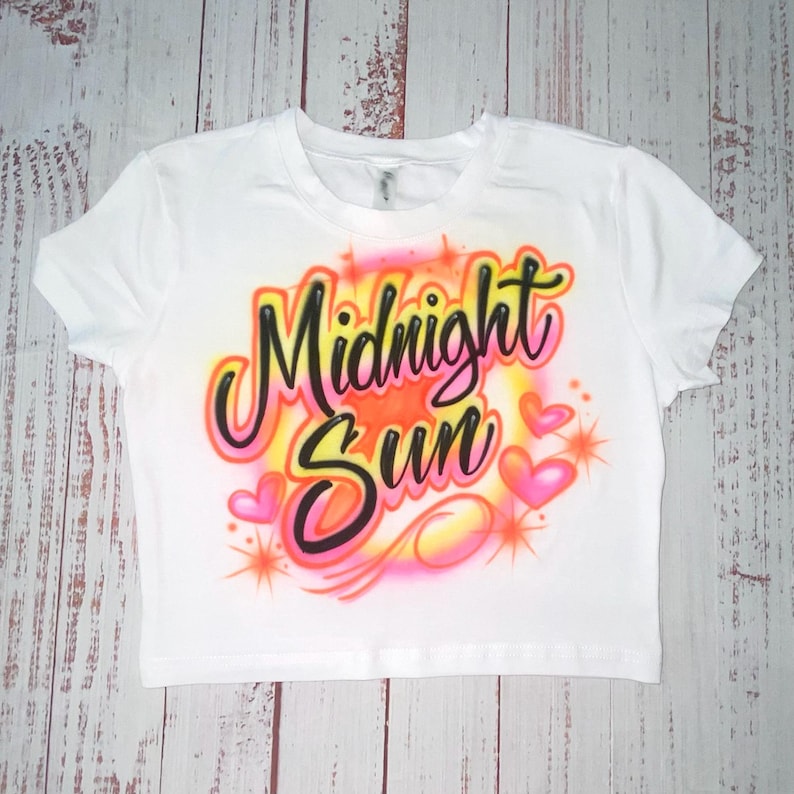 May include: White crop top with the words "Midnight Sun" in a colorful airbrushed design. The design includes pink hearts and orange starbursts, creating a vibrant and eye-catching graphic.