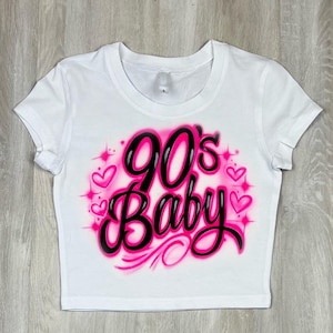 May include: White crop top with the text "90's Baby" in black and pink, surrounded by pink hearts and sparkles. The shirt has short sleeves and a crew neck.