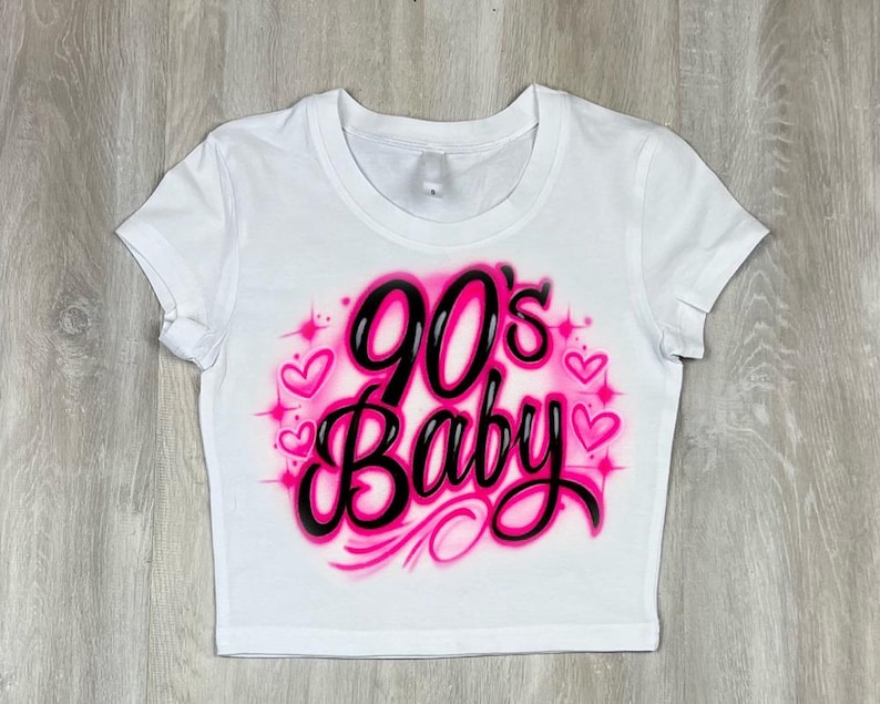 May include: White crop top with the text "90's Baby" in black and pink, surrounded by pink hearts and sparkles. The shirt has short sleeves and a crew neck.