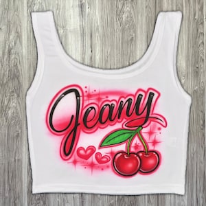 May include: White crop top with the name "Jeany" in a black and pink airbrushed font. Features two red cherries with green leaves and pink hearts. A fun, casual top.