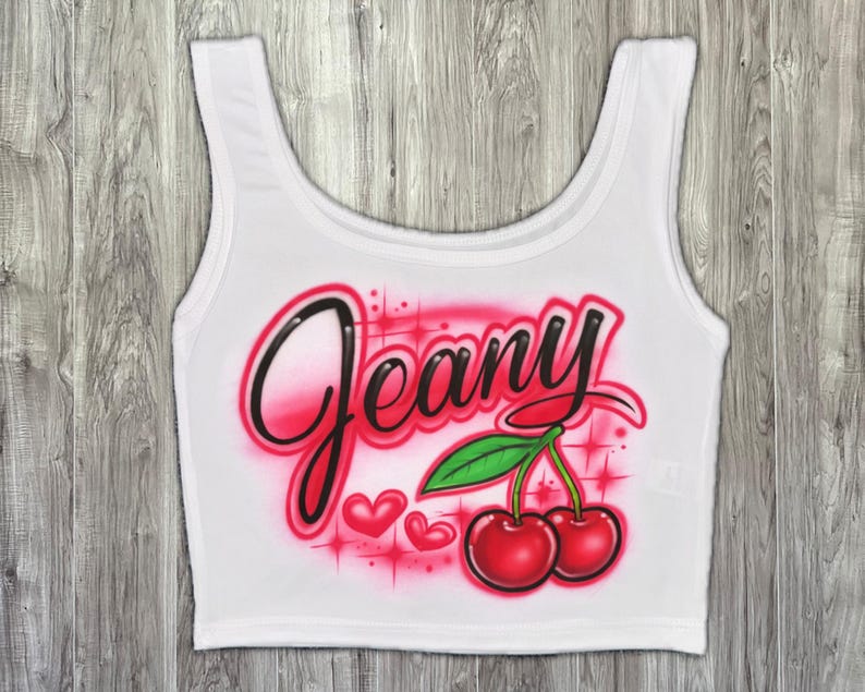 May include: White crop top with the name "Jeany" in a black and pink airbrushed font. Features two red cherries with green leaves and pink hearts. A fun, casual top.