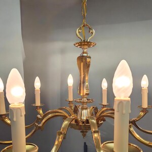 May include: A gold-toned brass chandelier with eight candle-style light bulbs. The chandelier has a decorative, ornate design with curved arms and a central finial.