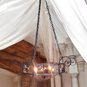 May include: A black wrought iron chandelier with a geometric design and ornate details hangs from a chain. The chandelier has multiple light bulbs that are turned on, casting a warm glow.