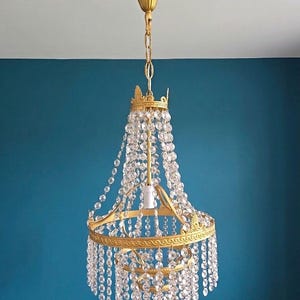 May include: A gold-toned metal chandelier featuring two circular tiers and a central rod. The fixture is adorned with numerous clear, faceted glass crystal beads arranged in cascading vertical strands that hang from the frame.