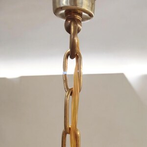 May include: Close-up of a gold-toned chain hanging from a ceiling fixture. The chain is made of metal links and has a decorative, braided cord running through it.