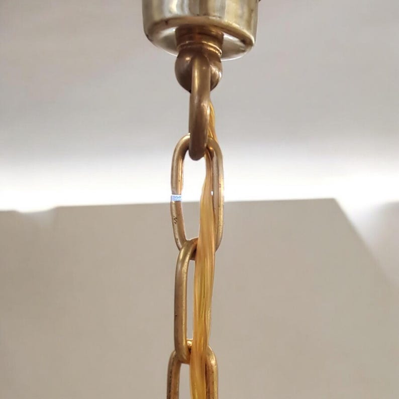 May include: Close-up of a gold-toned chain hanging from a ceiling fixture. The chain is made of metal links and has a decorative, braided cord running through it.