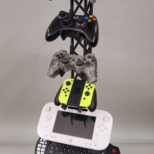 May include: A black metal stand with a variety of video game controllers, including a Nintendo Switch, a Wii U, a GameCube, and a PlayStation 3 controller. The stand also has a black keyboard and a black mouse.