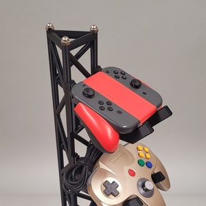 May include: A black metal stand with four different video game controllers. The controllers are a Nintendo Switch Joy-Con, a Nintendo 64 controller, a black Xbox controller, and a blue Playstation controller. The stand has a black base with the word "Fang" printed on it.
