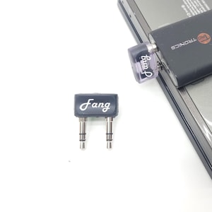 Puede incluir: Una consola de juegos portátil Nintendo 3DS negra y gris con un transmisor Bluetooth TRONICS negro conectado a la toma de auriculares. El transmisor tiene un pequeño botón redondo plateado con un símbolo 'G' en él. También se ve un pequeño adaptador morado transparente con la palabra 'Fang' impresa.