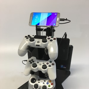 May include: A black stand with a star-shaped base holds three video game controllers and a smartphone. The stand is labeled "Fang".
