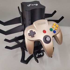 Puede incluir: Un mando de Nintendo 64 dorado se encuentra sobre un soporte negro con forma de araña con la palabra "Fang" impresa en el soporte.