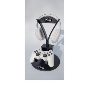 May include: A black stand with a white gaming headset and a white game controller. The stand has the word "Fang" printed on it.