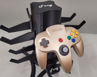 Puede incluir: Un mando de Nintendo 64 dorado se encuentra sobre un soporte negro con forma de araña con la palabra "Fang" impresa en el soporte.