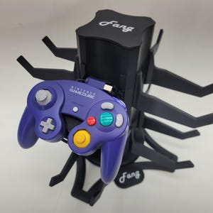 Puede incluir: Un soporte negro para mandos de GameCube con la palabra "Fang" impresa en él. El soporte tiene seis brazos que sujetan los mandos. Un mando de GameCube morado está en uno de los brazos. El mando tiene las palabras "Nintendo GameCube" impresas en él.