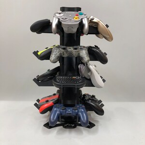 May include: A black metal stand with six slots for video game controllers. The stand has a black base with the word "Fang" printed on it. The stand is holding a variety of controllers, including a silver GameCube controller, a black and green camouflage controller, a white Wii remote, a red and black controller, and a blue and black controller.