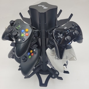 Könnte beinhalten: Ein schwarzer Plastik-Videospiel-Controller-Ständer mit dem Wort "Fang" oben. Der Ständer hält mehrere Controller, darunter Xbox, Wii und andere Controller.