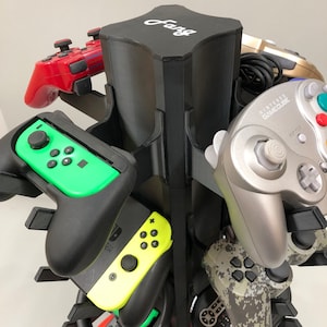 May include: A black plastic stand with slots to hold multiple video game controllers. The stand has the word "Fang" printed on the top. The controllers include a red and black Nintendo Switch controller, a green and black Nintendo Switch controller, a silver Nintendo GameCube controller, a camouflage-patterned controller, and a gold and black controller.