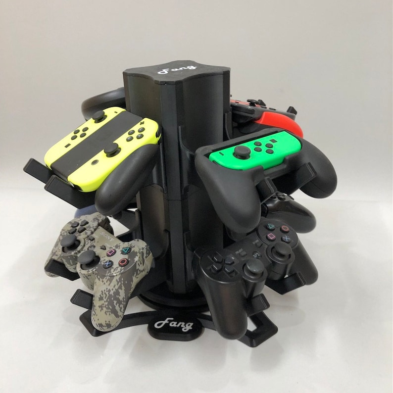 May include: A black rotating stand with the word "Fang" printed on it. The stand holds multiple video game controllers, including a yellow and black Nintendo Switch controller, a green and black Nintendo Switch controller, and two black PlayStation controllers.