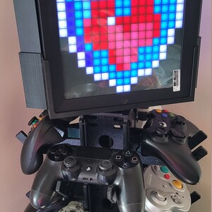May include: A black stand with a small screen displaying a pixelated heart in red and blue. The stand holds four different video game controllers, including a PlayStation 4 controller, a Nintendo GameCube controller, and two Xbox controllers.