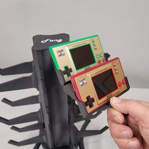 May include: A black plastic stand with the word "Fang" printed on it holds two vintage Nintendo Game & Watch handheld video game consoles. The Game & Watch consoles are gold, red, and green. The console on the left is "The Legend of Zelda" and the console on the right is "Super Mario Bros."