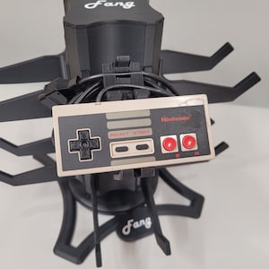 May include: A black plastic stand with the word "Fang" printed on it. The stand has multiple arms that hold a gray and white Nintendo Entertainment System controller. The controller has the word "Nintendo" printed on it.