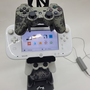 May include: A white Nintendo Wii U console with a camouflage-patterned Sony Playstation 3 controller attached to a black stand. The console is displaying the Wii U menu with various icons. The stand also has a white Nintendo Wii U Pro Controller attached to it.