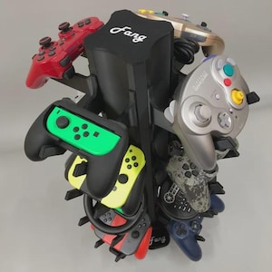 May include: A black vertical gaming controller stand with the word Fang printed in white on the top and base. The stand holds multiple controllers, including a red one, a silver GameCube controller, a neon green and black one, and others.