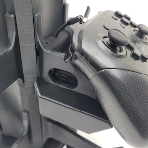 May include: A black video game controller with textured grips and buttons is mounted on a black stand. The controller is connected to the stand with a black cable.