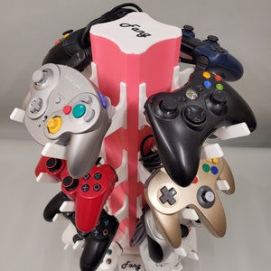 May include: A pink and white video game controller stand with the word "Fang" printed on it. The stand holds nine different video game controllers, including a Nintendo 64 controller, a GameCube controller, a PlayStation 3 controller, and an Xbox 360 controller.