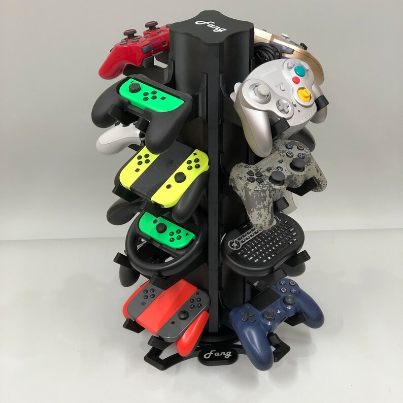 May include: A black rotating stand with the word "Fang" on it holds a variety of video game controllers, including a yellow and black Nintendo Switch controller, a green and black Nintendo Switch controller, a red and black Nintendo Switch controller, a blue and black PlayStation controller, a black and white GameCube controller, a black and white Wii controller, and a black and white GameCube controller.