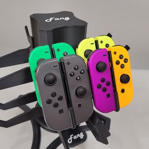 May include: A black stand with the word "Fang" printed on it holds five different colored Nintendo Switch Joy-Con controllers. The controllers are green, yellow, purple, and orange.