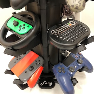 May include: A black rotating stand with multiple slots for storing video game controllers. The stand has a black base with the word "Fang" printed on it. The stand is holding a variety of controllers, including a green and black Nintendo Switch controller, a red and gray Nintendo Switch controller, a blue PlayStation 4 controller, and a black wireless keyboard with a trackball.