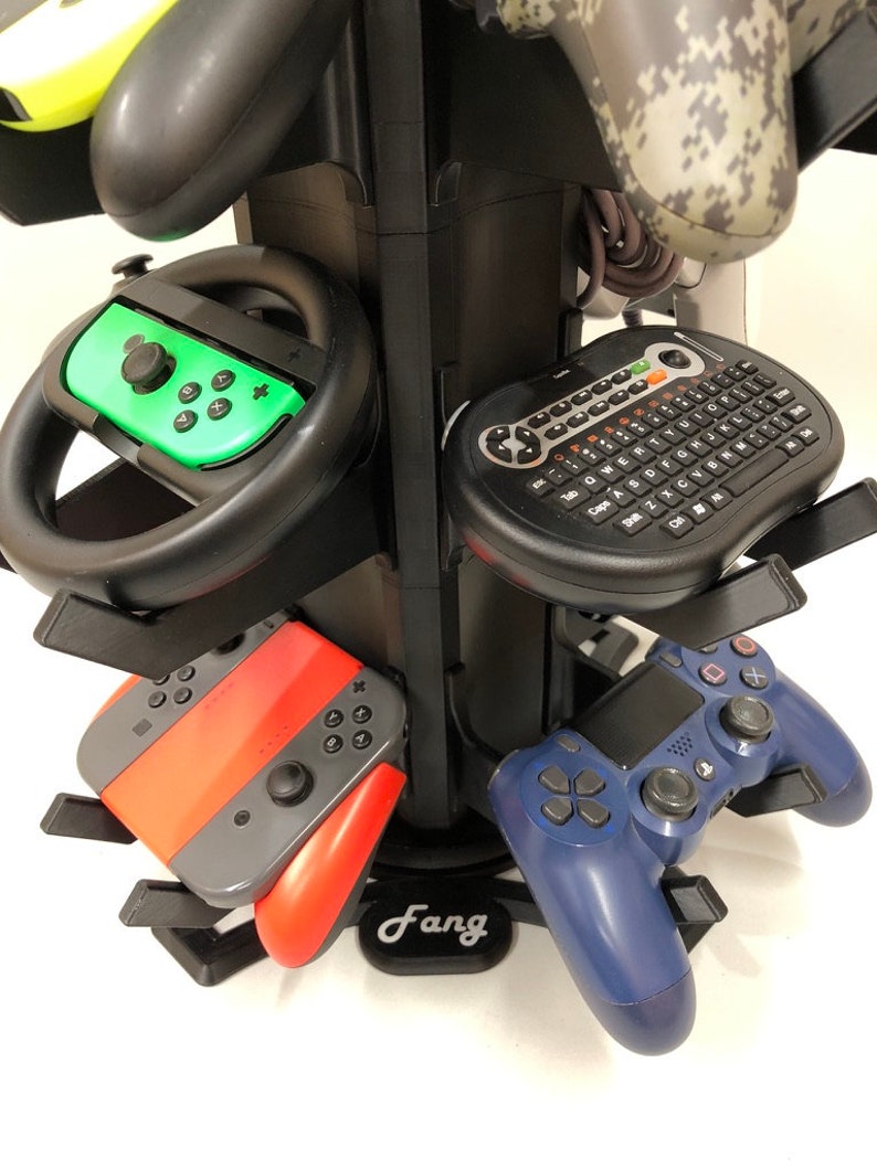 May include: A black rotating stand with multiple slots for storing video game controllers. The stand has a black base with the word "Fang" printed on it. The stand is holding a variety of controllers, including a green and black Nintendo Switch controller, a red and gray Nintendo Switch controller, a blue PlayStation 4 controller, and a black wireless keyboard with a trackball.