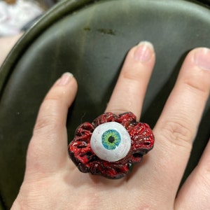 May include: A ring with a red fleshy design and a white eyeball with a green and blue iris. The ring is worn on a finger.