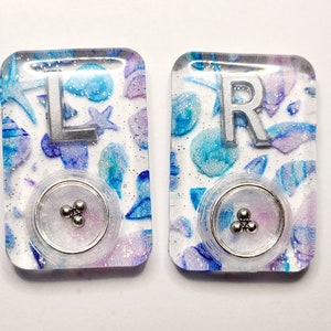 May include: Two clear resin keychains with a white background and blue and purple glitter. Each keychain has a silver letter, one says "L" and the other says "R". Each keychain has a clear circle with three silver balls inside.