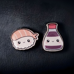May include: Two cute cartoon pins, one of a sushi roll with a pink salmon filling and the other of a soy sauce bottle. Both have smiling faces.