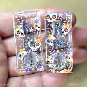 May include: Two rectangular resin earrings with a Day of the Dead theme. Each earring has a silver letter, "L" and "R", and the letters "RB". The earrings feature sugar skull designs, flowers, and a small shaker element with silver beads.