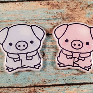 May include: Two pig-shaped resin charms, one light blue and one pink, with black outlines and cute cartoon faces. The charms are both wearing a jacket with a collar and have their arms crossed.