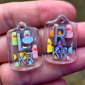 May include: Two clear, rectangular keychains with rounded tops. Each keychain features a letter and the letters "KR" in silver. The keychains are decorated with colorful duck illustrations. The keychain on the left has the letter "L". The keychain on the right has the letter "R".