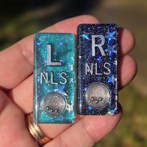 May include: Two rectangular resin markers, one teal and one dark purple, both embedded with holographic star glitter. Each marker features the silver letters L or R above the text NLS and a small circular clear compartment containing three tiny metal balls.