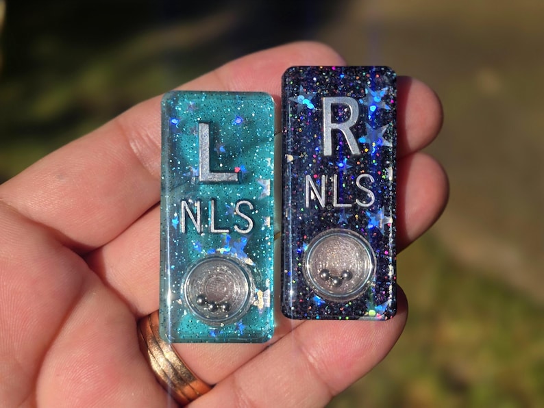May include: Two rectangular resin markers, one teal and one dark purple, both embedded with holographic star glitter. Each marker features the silver letters L or R above the text NLS and a small circular clear compartment containing three tiny metal balls.