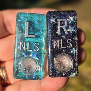 May include: Two rectangular resin charms, one teal and one black, both filled with holographic star glitter. Each charm features the silver letters L or R above the letters NLS, and a small circular clear window containing three tiny metal ball bearings.