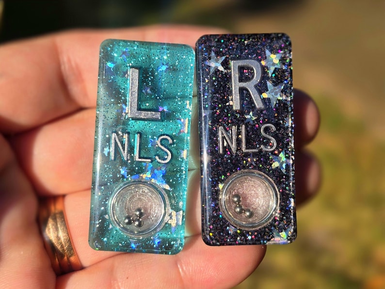 May include: Two rectangular resin charms, one teal and one black, both filled with holographic star glitter. Each charm features the silver letters L or R above the letters NLS, and a small circular clear window containing three tiny metal ball bearings.