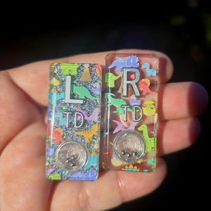 May include: Two rectangular resin charms featuring colorful dinosaur patterns and silver-toned letters. The left charm displays the letters L and TD with silver glitter, while the right charm shows R and TD. Each piece includes a small circular shaker element.