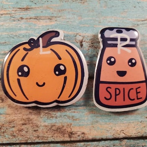 May include: Two cute cartoon-style pins, one of an orange pumpkin with a happy face and the other of a red spice bottle with a happy face. Both pins have glitter accents and say "SPICE" on the bottle.