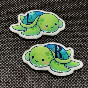 May include: Two green and blue turtle-shaped resin ornaments with a letter on each. The first turtle has the letter "L" and the second turtle has the letter "R".